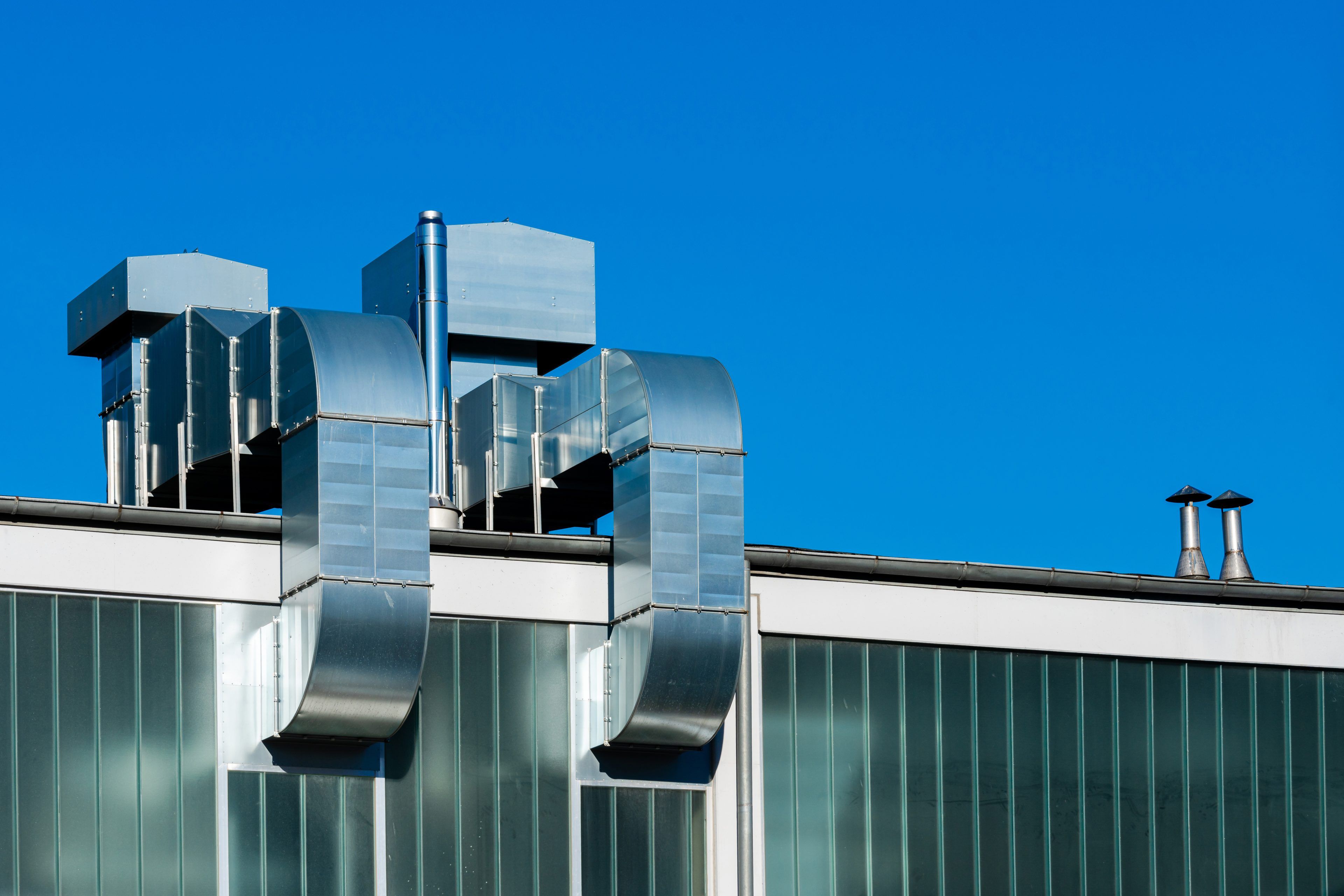 Metal ventilation pipes and ducts are seen on a modern building against a clear blue sky. The structure features glass panels, showcasing contemporary architectural design.