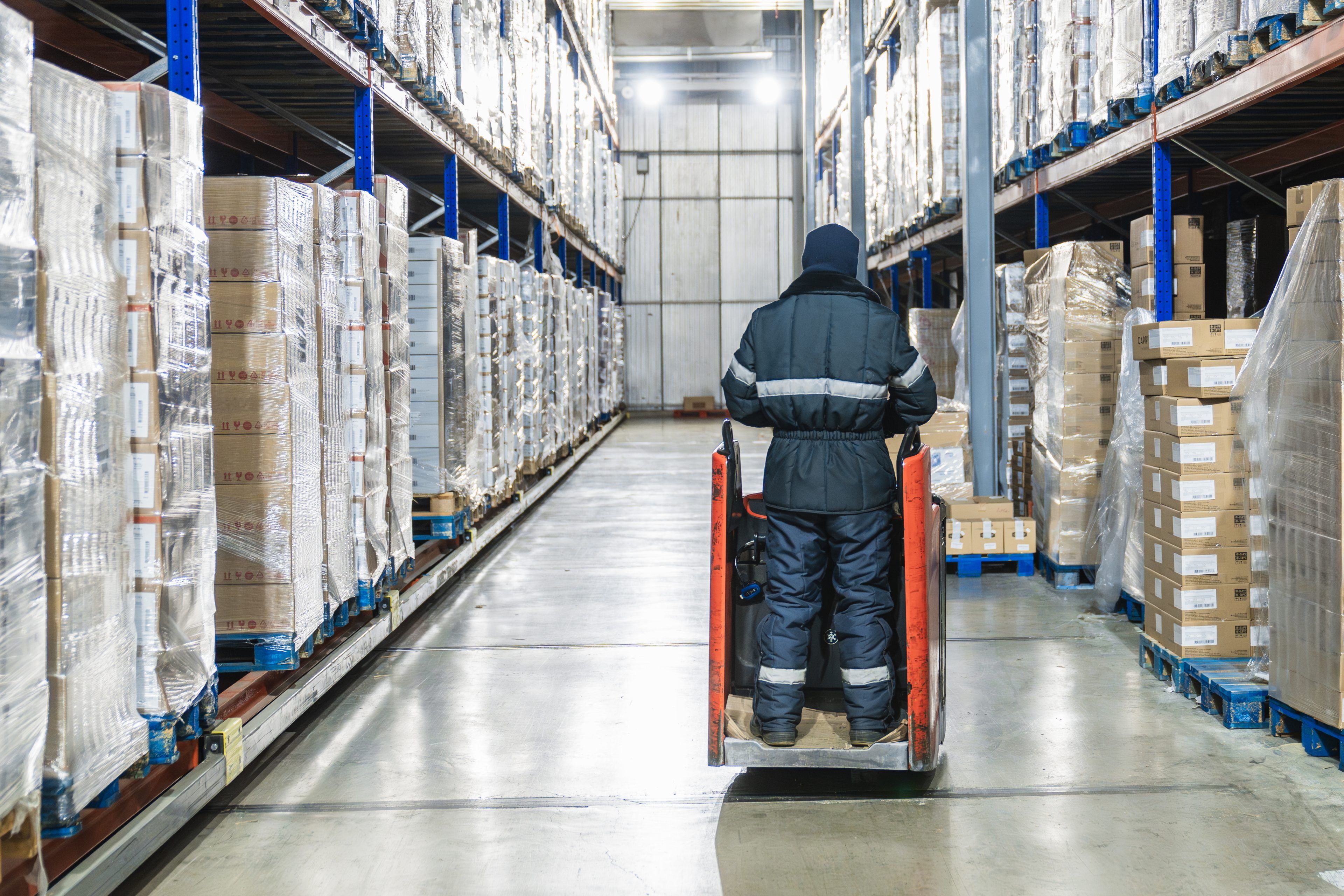 Worker in warm clothing moving pallets of goods with a forklift in a large low temperature cold storage facility